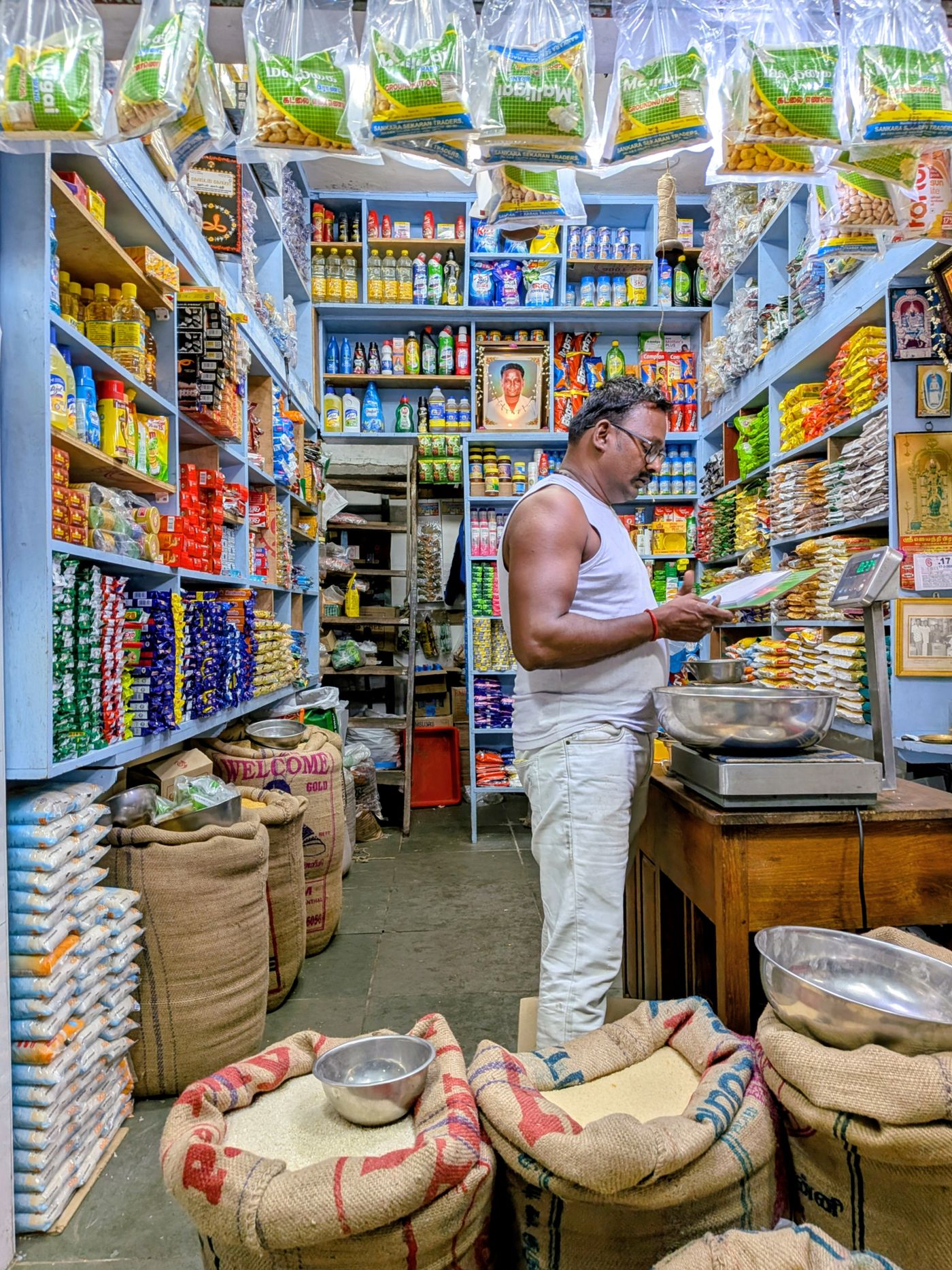 A kirana store owner behind a counter stacked with grocery packets